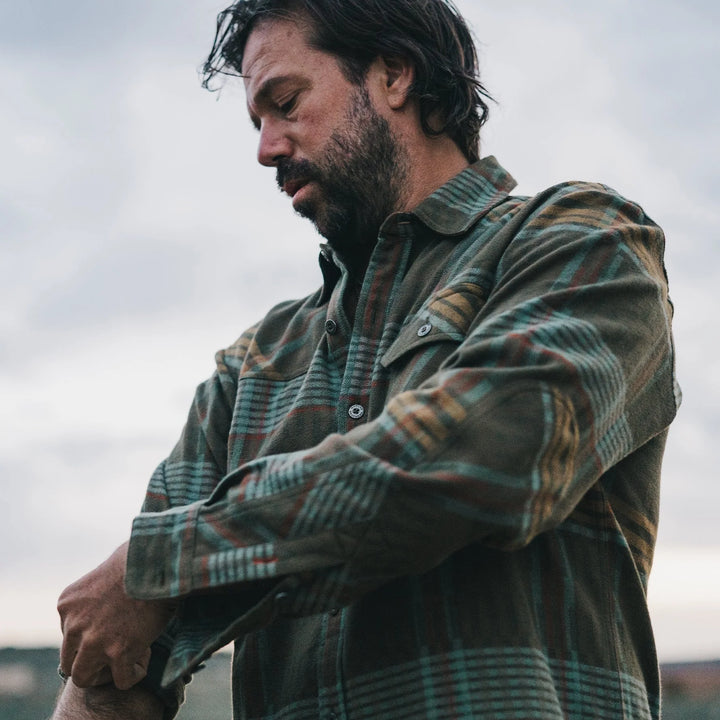 A long-sleeved plaid flannel shirt with a button-through flap pocket on the left chest, reinforced elbow patches, and custom metal buttons.