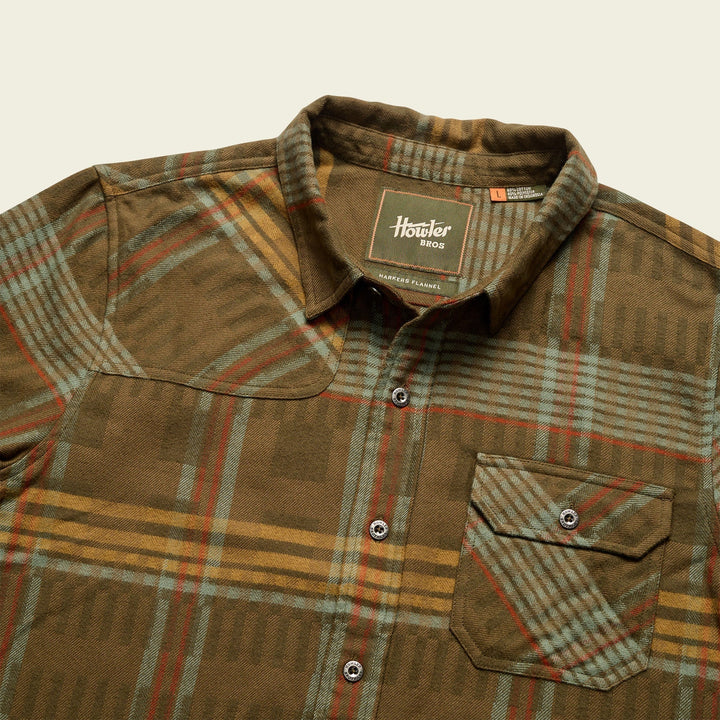 A long-sleeved plaid flannel shirt with a button-through flap pocket on the left chest, reinforced elbow patches, and custom metal buttons.