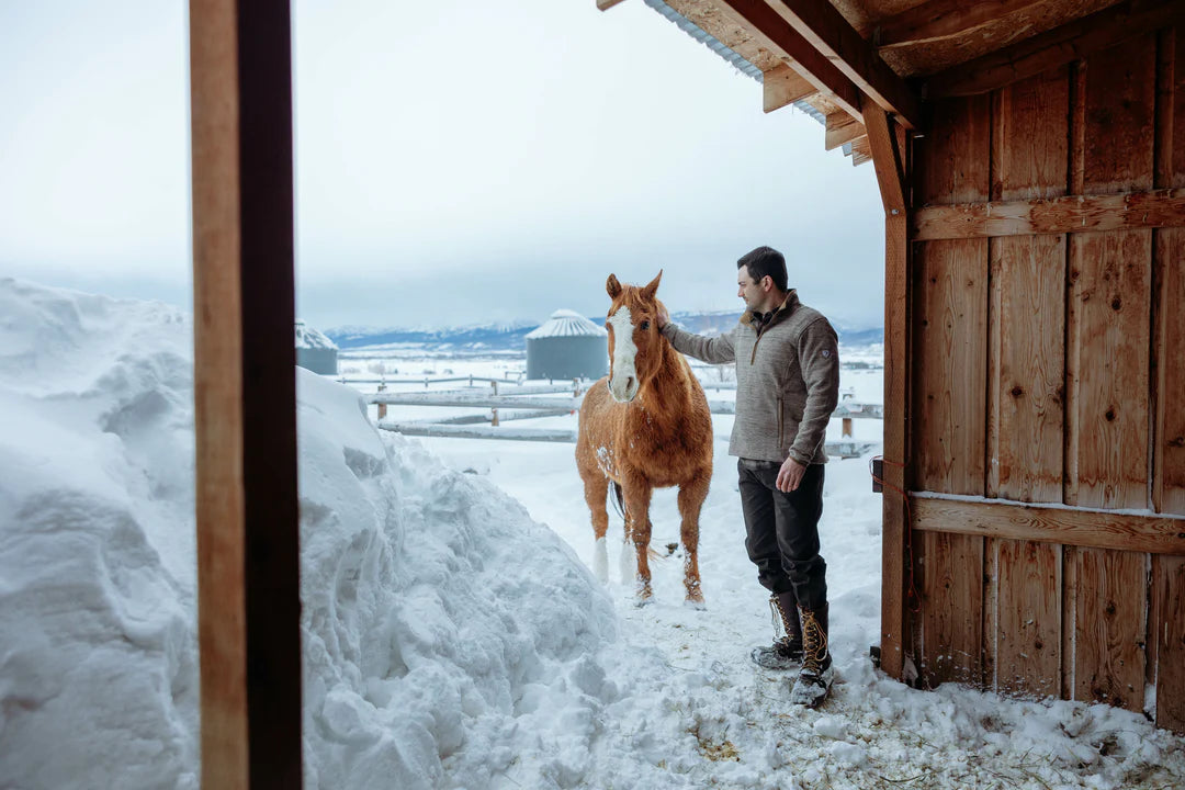 A man wearing Kuhl Rydr pants in the snow petting a horse near a barn