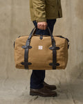Person holding a tan duffel bag with black straps against a beige background