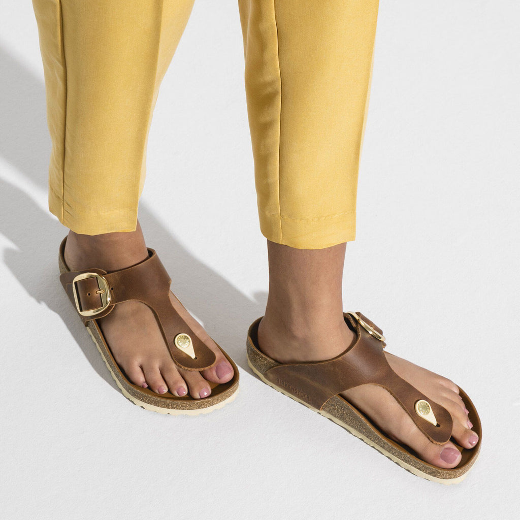 Brown sandals with gold buckles worn by a person in yellow pants on a light gray background
