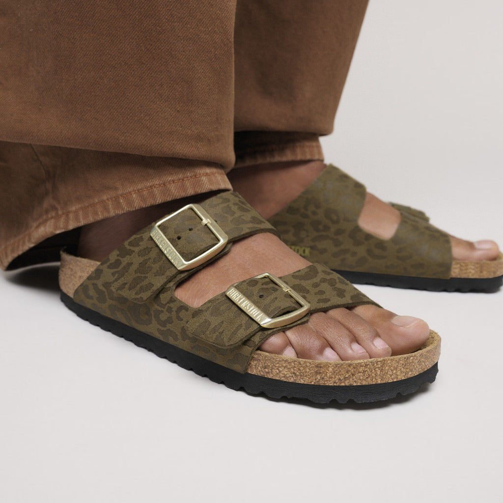 Brown sandals with gold buckles worn by a person in brown pants on a light gray background
