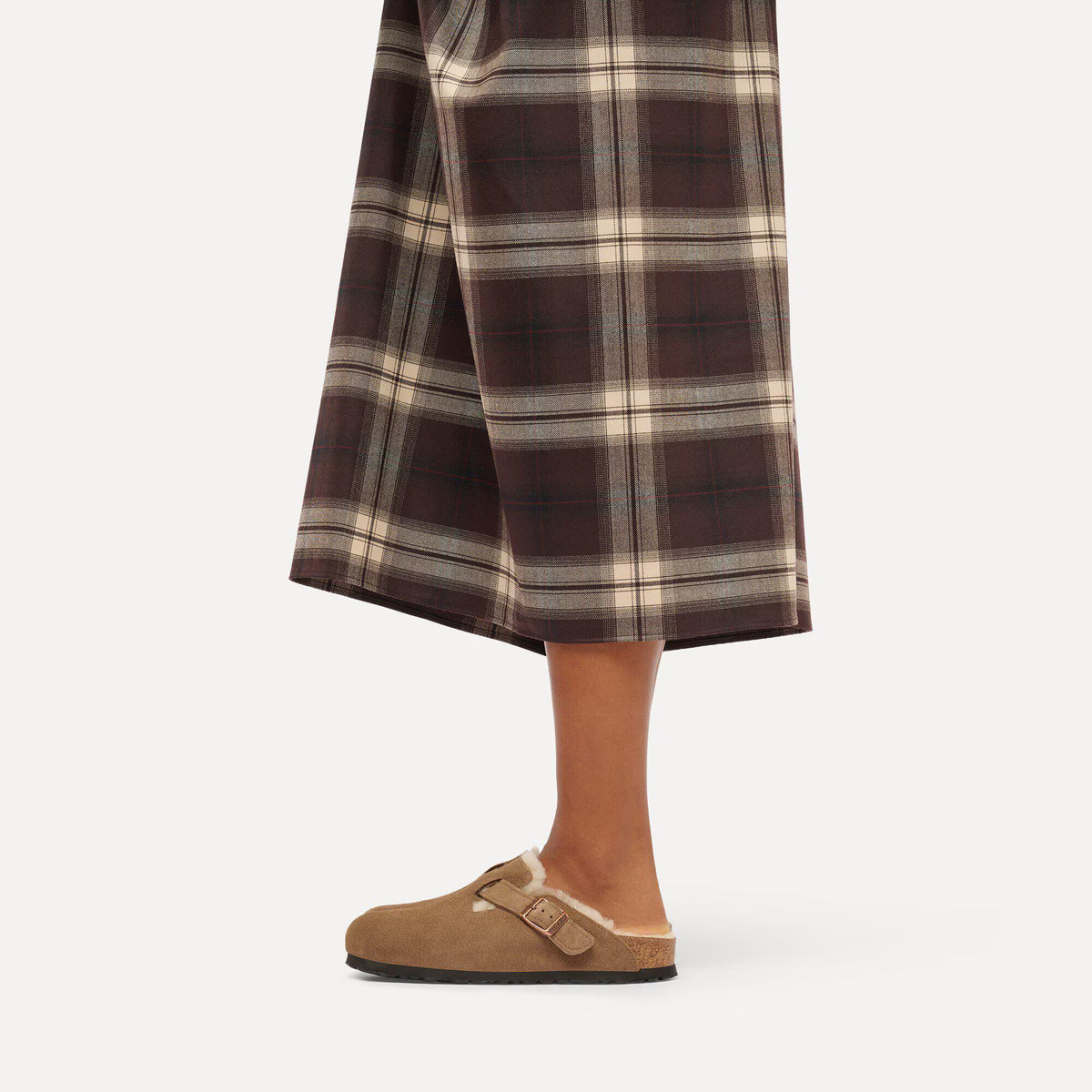 A person wearing brown and beige plaid wide-leg cropped pants with tan slip-on clogs, shown from the waist down against a light gray studio background.