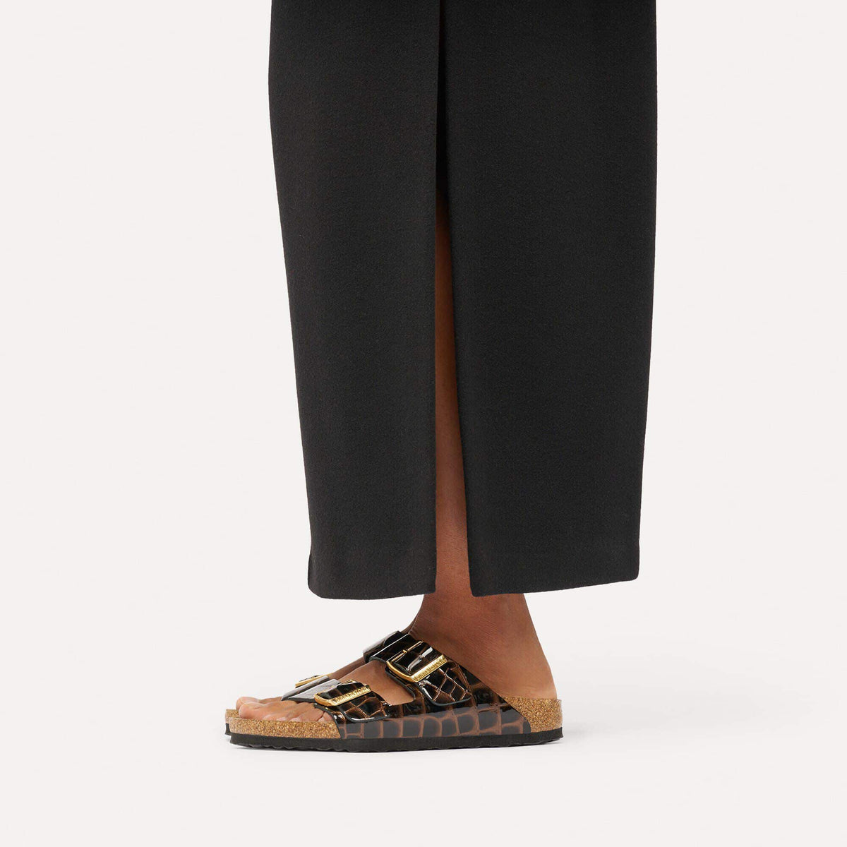 A close-up of a person's foot wearing a pair of black wide-leg trousers with a front slit and brown and gold double-strap slide sandals with a cork midsole, set against a plain light background.