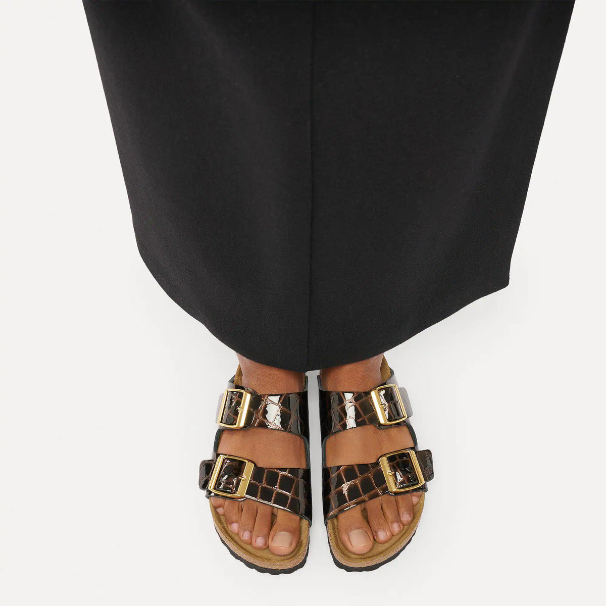 Brown sandals with gold buckles worn with black pants on a white background