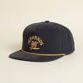 A dark gray corduroy baseball cap with a gold embroidered logo reading 'HERMANOS' and a bird emblem on the front, plus a gold stitched line along the brim, shown on a plain white background.