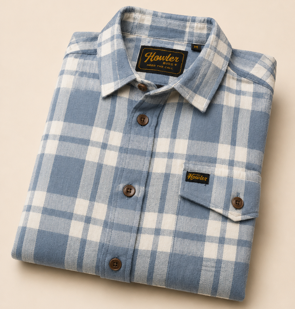 A folded blue and white plaid button-down shirt with a chest pocket, displayed on a warm beige background. The brand 'Howler' is visible on the collar label and the chest pocket tag.