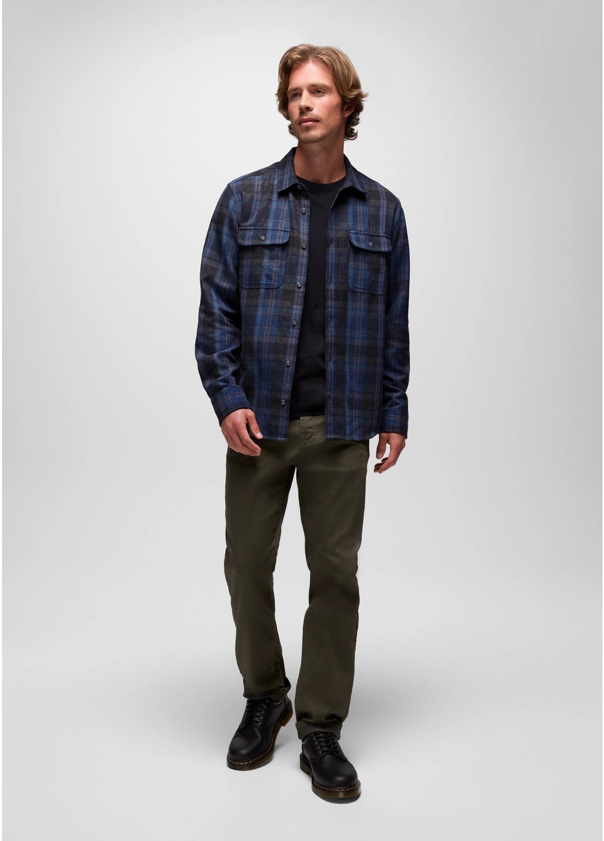 Man wearing a blue plaid shirt over a black shirt, green pants, and black shoes on a white background