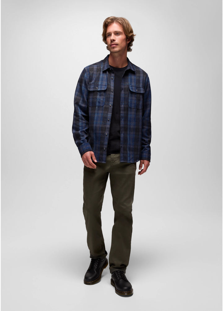 Man wearing a blue plaid shirt over a black shirt, green pants, and black shoes on a white background