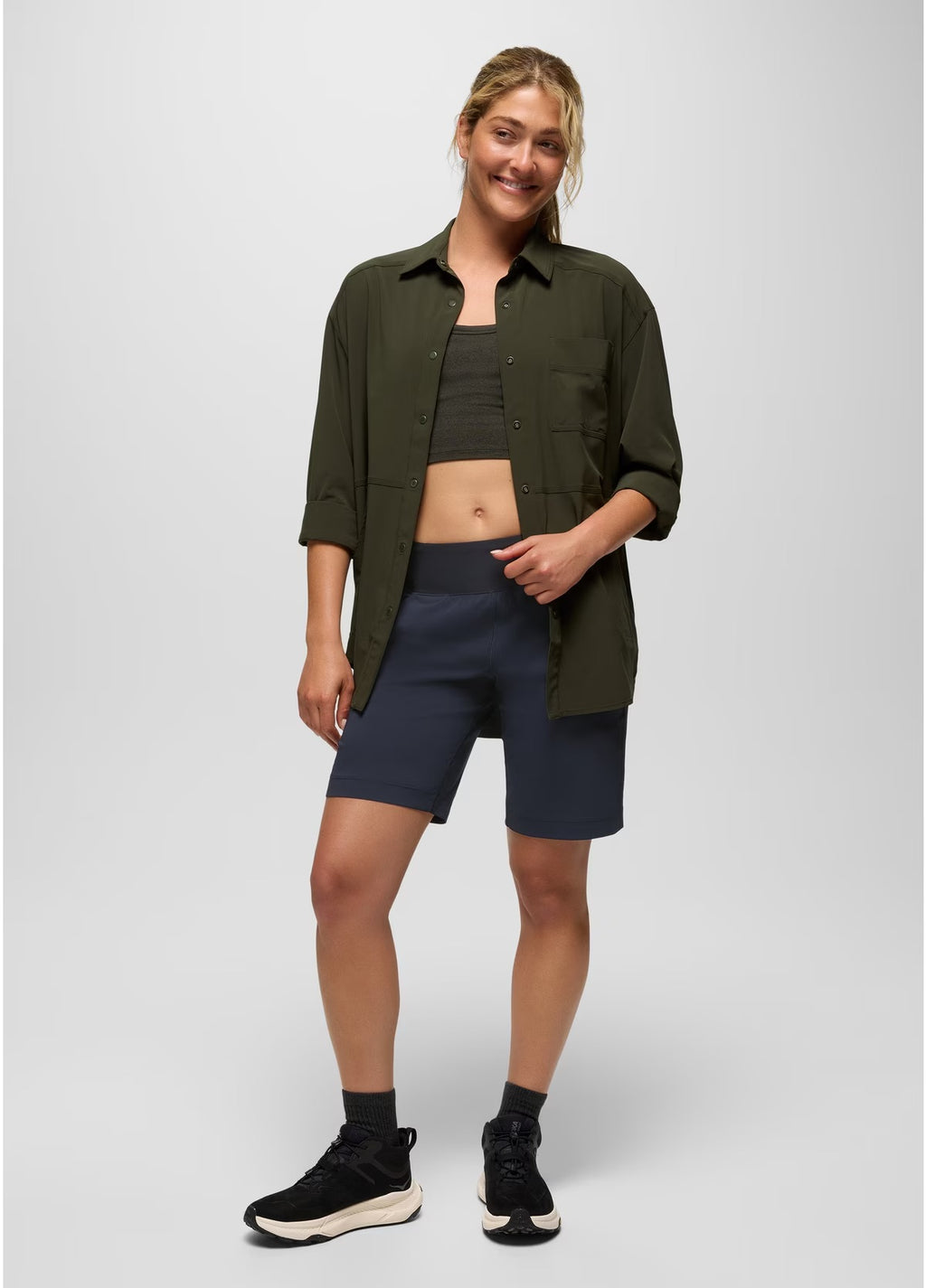 Woman wearing an olive green shirt, black top, and navy shorts on a white background