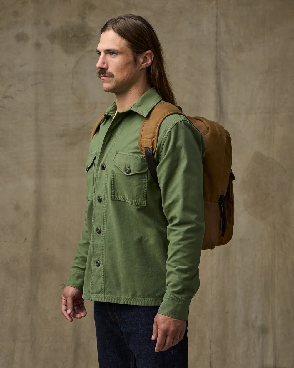 A man with long brown hair and a mustache is shown in profile wearing an olive green button-up shirt and a brown backpack, standing against a textured concrete wall.
