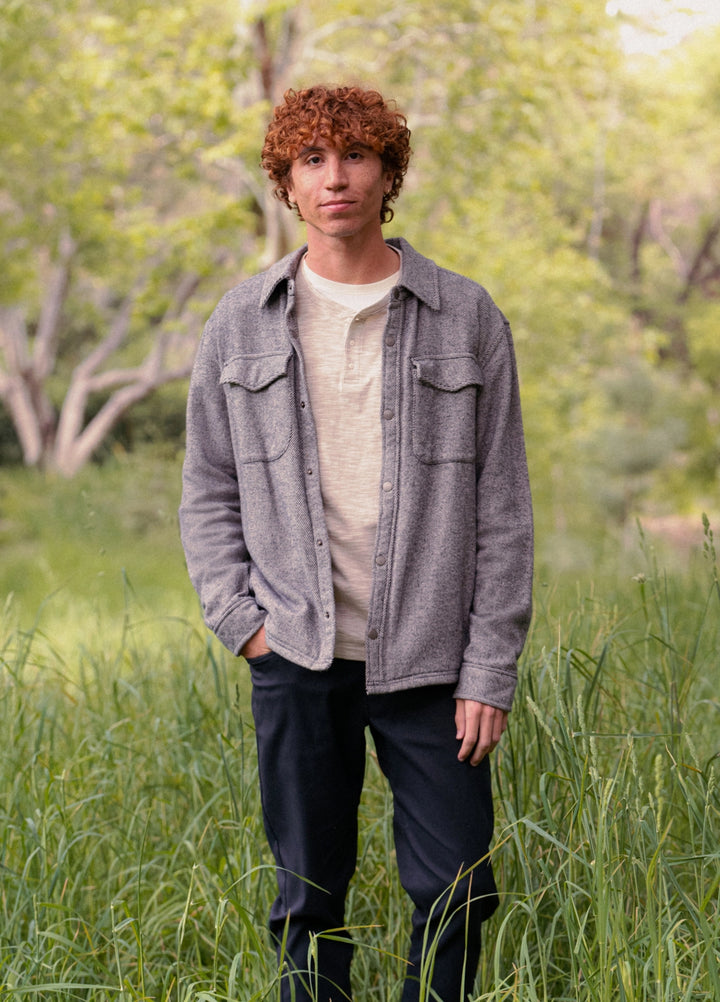 Person wearing a gray jacket and dark pants standing in a grassy field with trees in the background