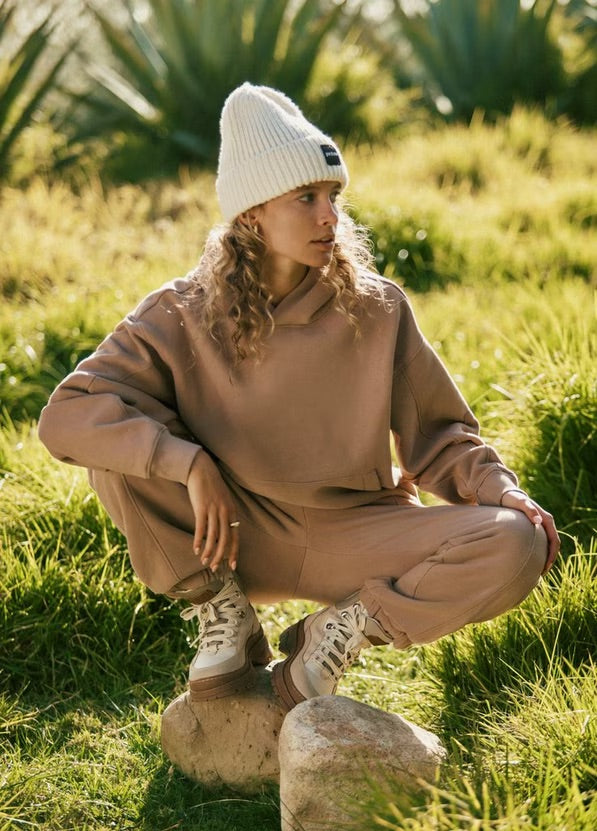 Person in beige outfit and white beanie sitting on a rock in a grassy field