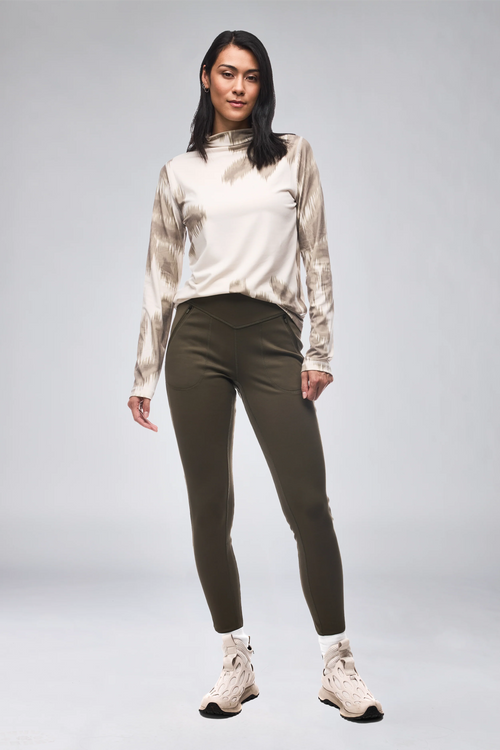 A woman modeling a long-sleeve athletic top with a beige and taupe tie-dye pattern and olive green leggings, standing against a light gray studio background.