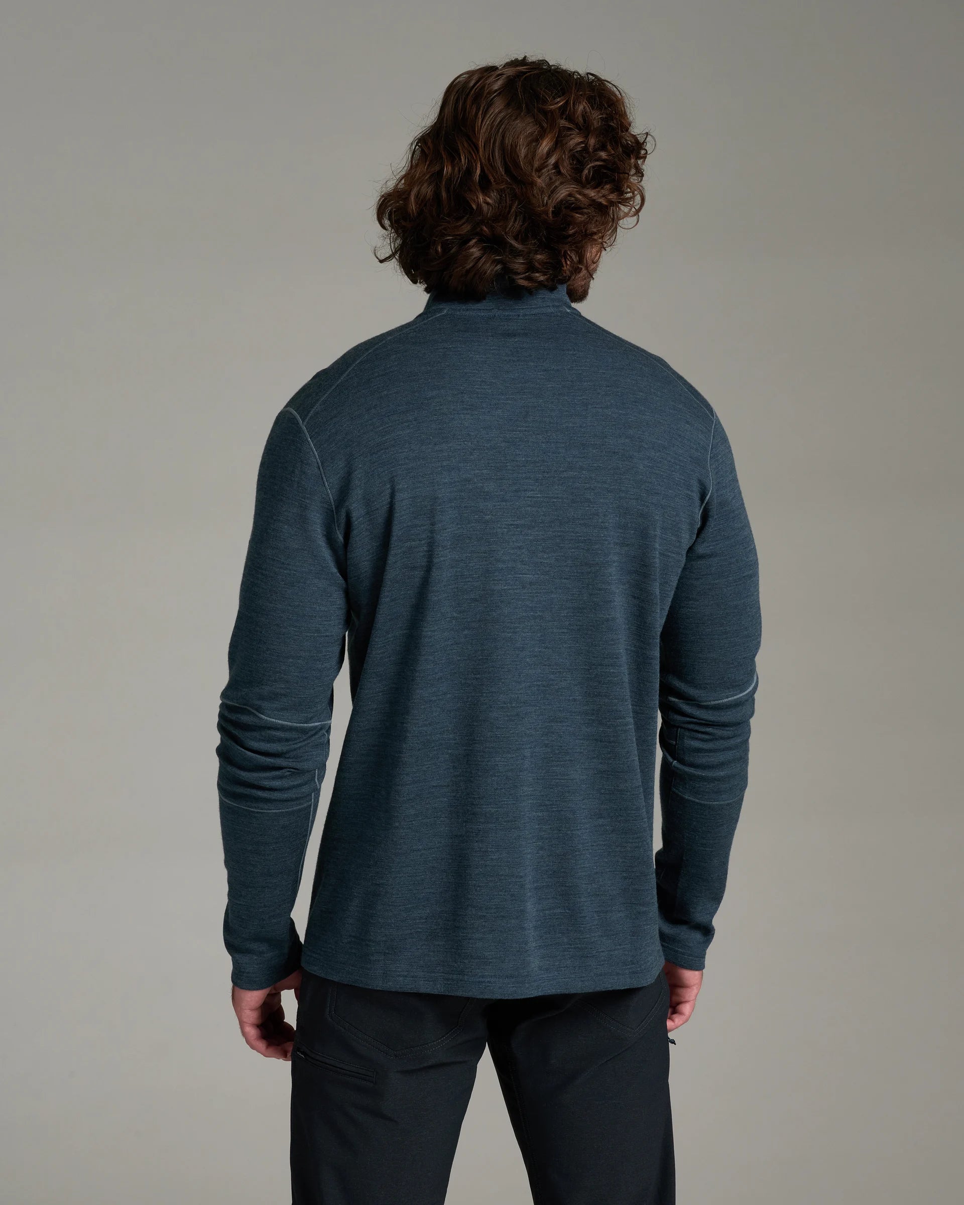 A man viewed from behind wearing a dark slate blue long-sleeve knit top and dark pants, standing against a neutral gray studio background.