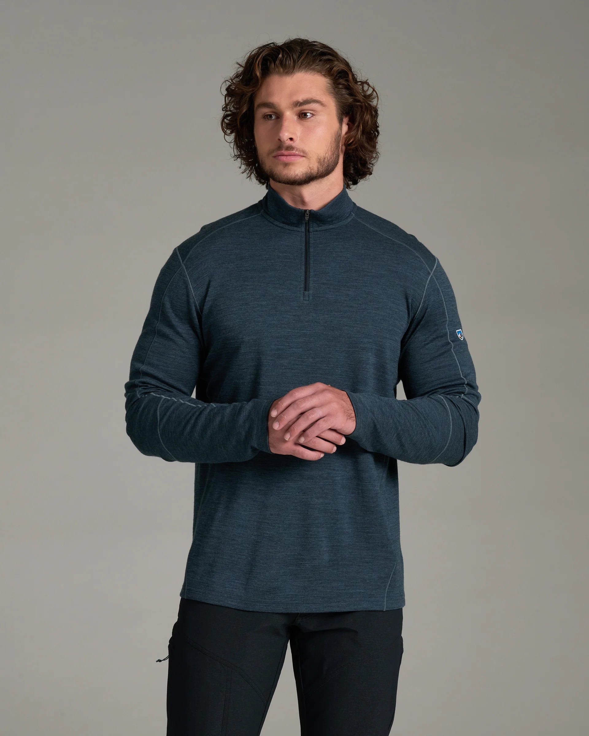 A man viewed from behind wearing a dark slate blue long-sleeve knit top and dark pants, standing against a neutral gray studio background.