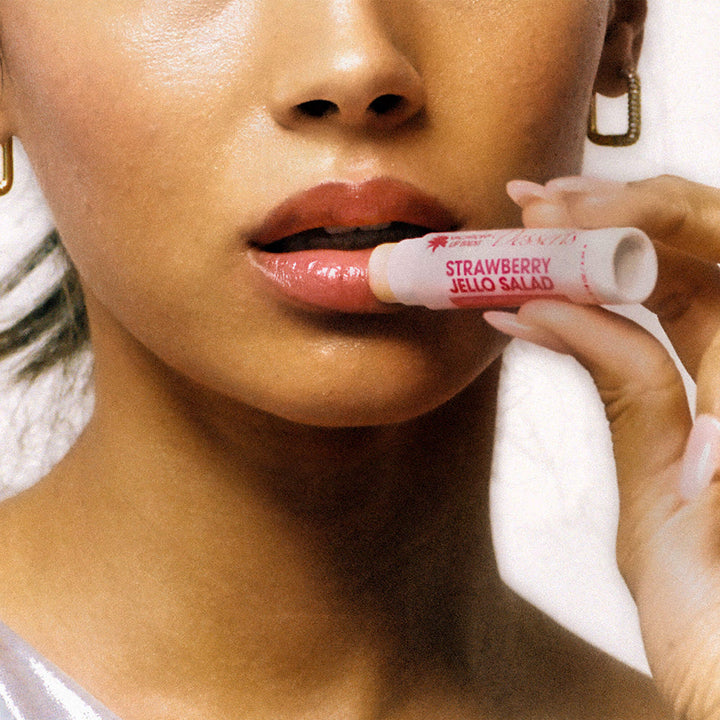 Person holding a tube of 'Strawberry Jello Salad' lip balm close to their mouth.
