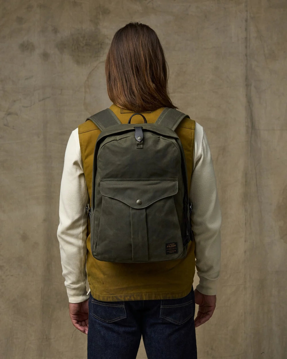 An olive green canvas backpack with a front flap pocket, shown from the back on a model wearing a mustard yellow vest over a cream long-sleeve shirt and blue jeans, set against a textured concrete wall.