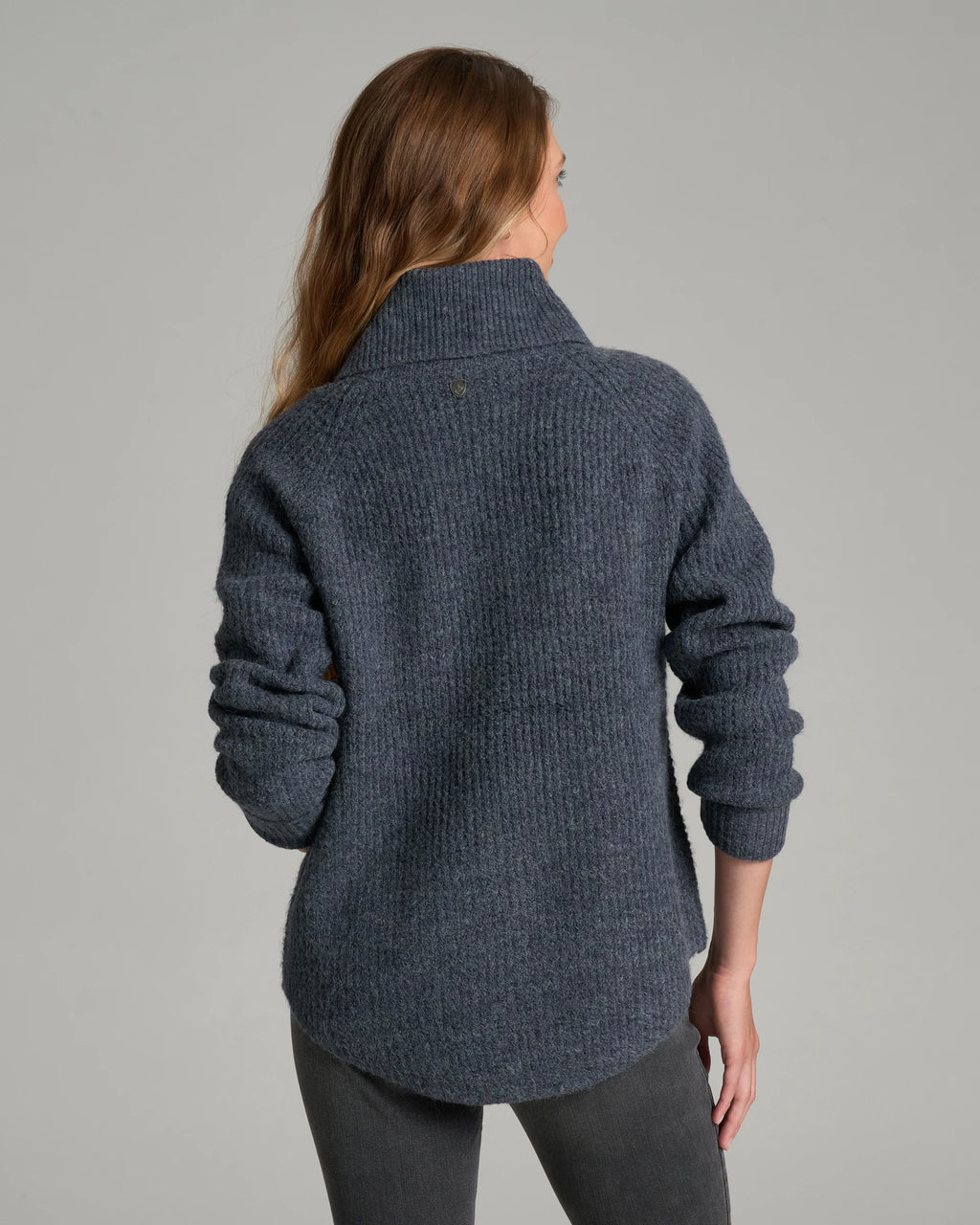 A woman viewed from the back wearing a dark blue-gray ribbed knit turtleneck sweater and dark gray pants, photographed against a plain light gray studio background.