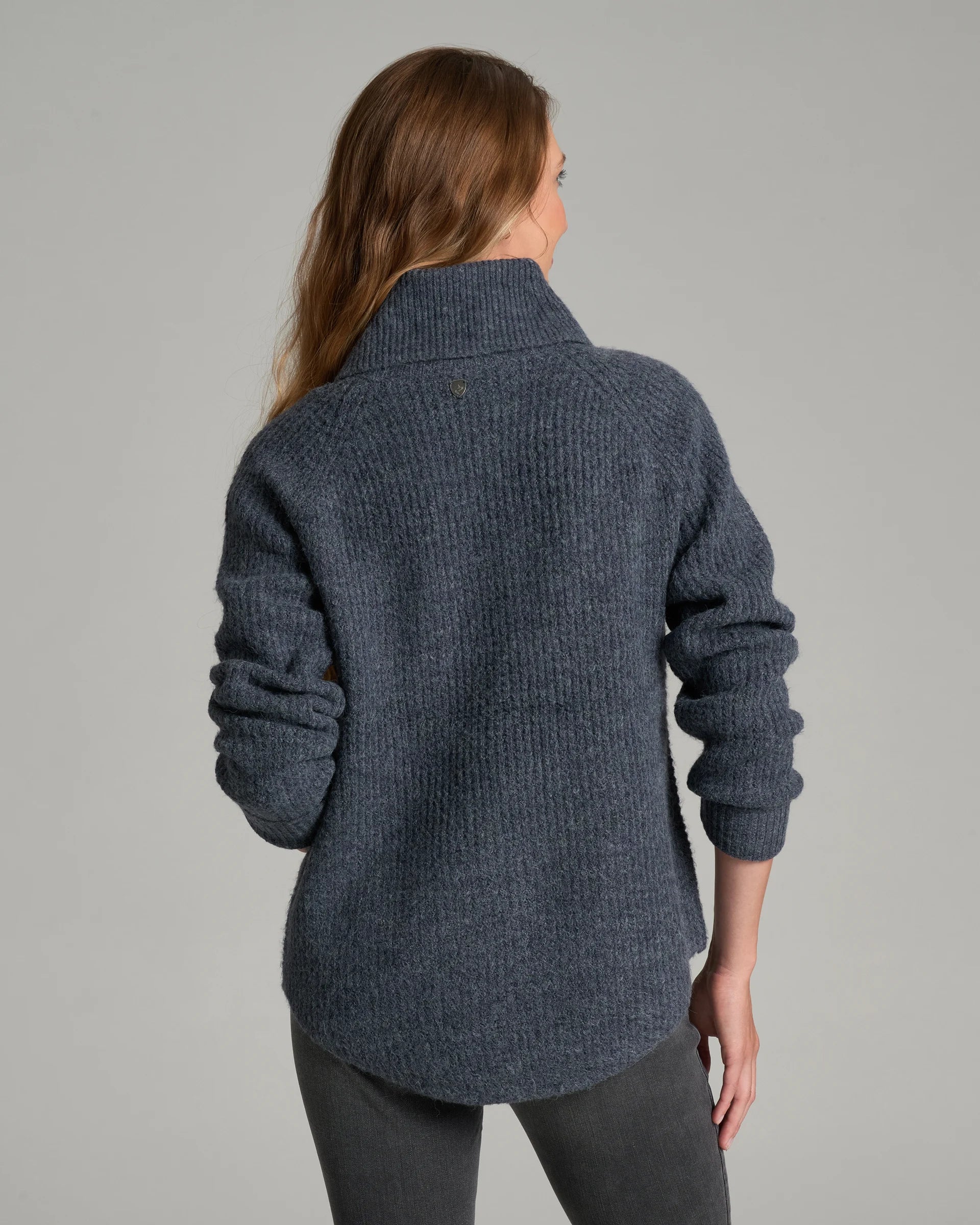 A woman viewed from the back wearing a dark blue-gray ribbed knit turtleneck sweater and dark gray pants, photographed against a plain light gray studio background.