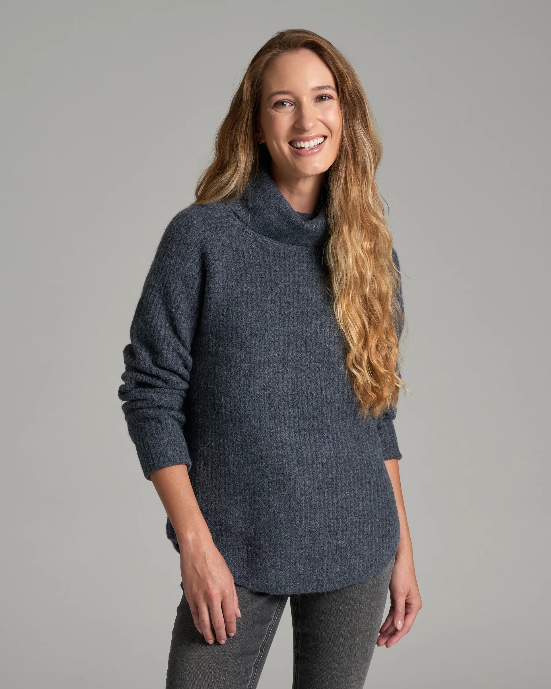 A woman with long blonde hair wearing a dark slate blue ribbed knit turtleneck sweater and gray pants, standing against a solid gray studio background.