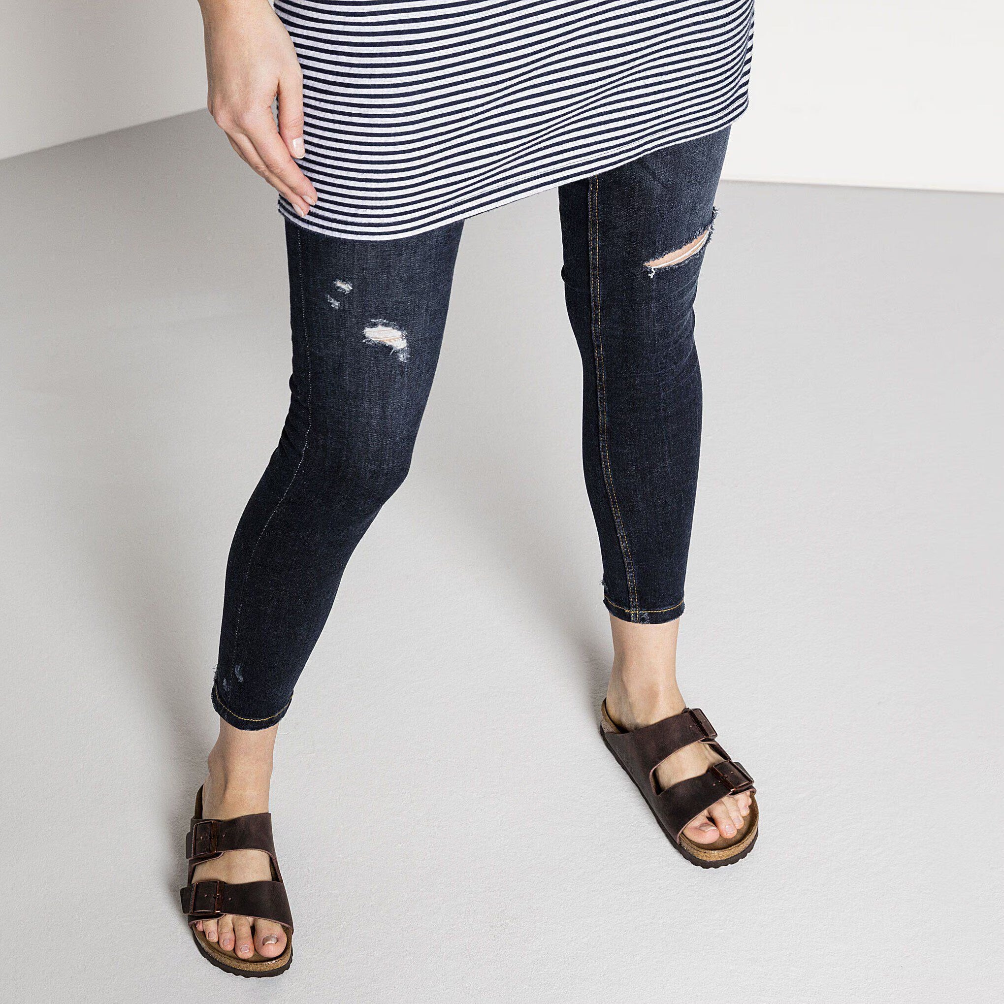 Person wearing distressed jeans, a striped shirt, and brown sandals on a plain background
