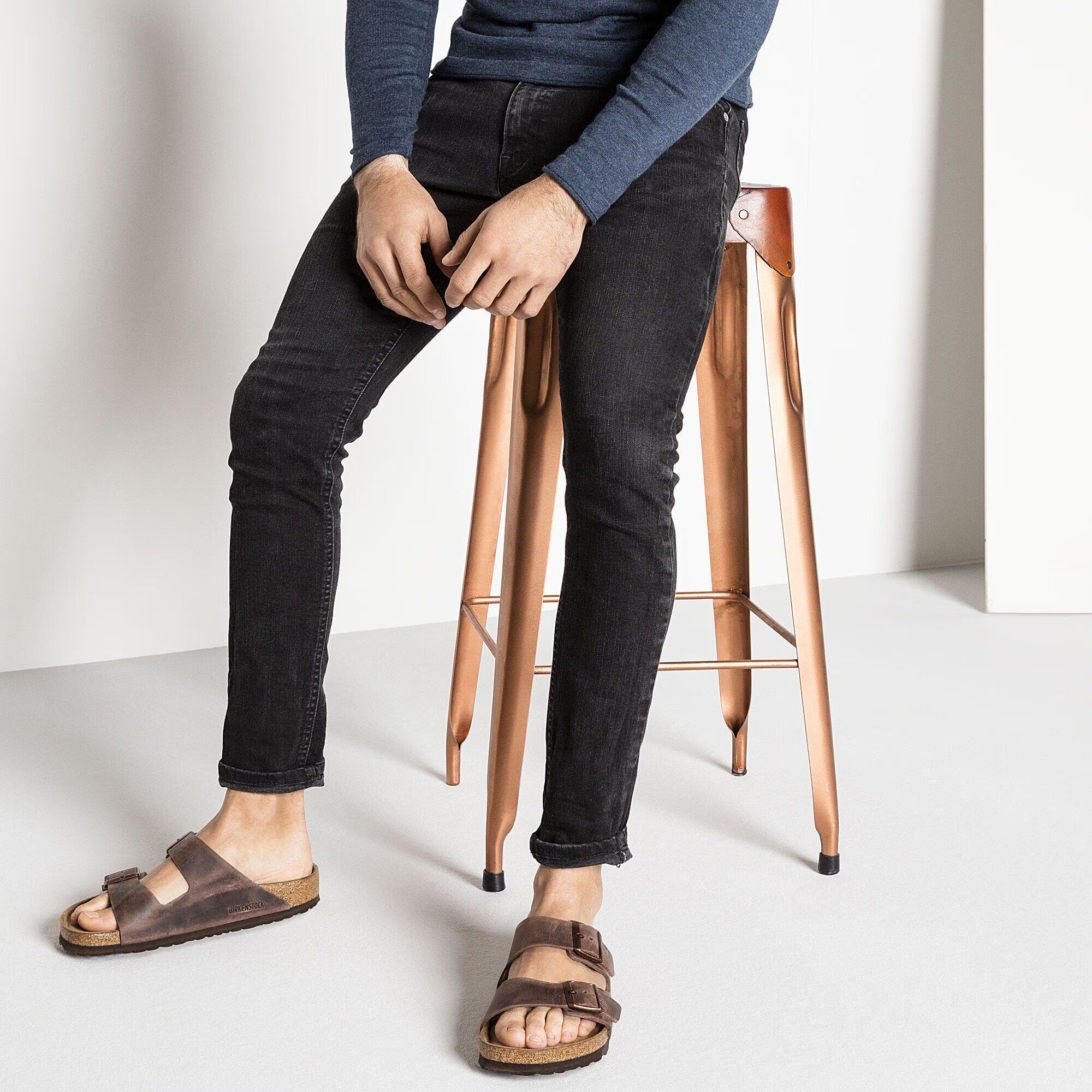 Person wearing black jeans and brown sandals sitting on a copper stool against a white background