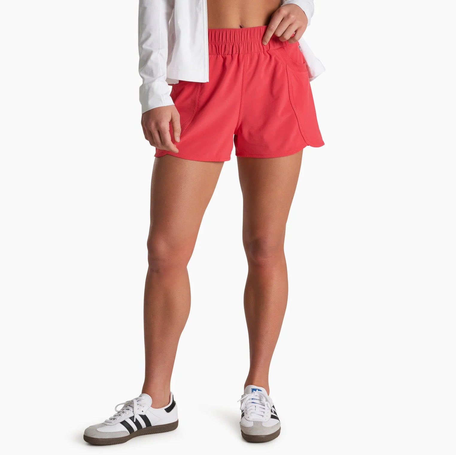 A model wearing bright coral pink athletic shorts, styled with a white long-sleeve shirt and white sneakers with black stripes, against a plain white background.