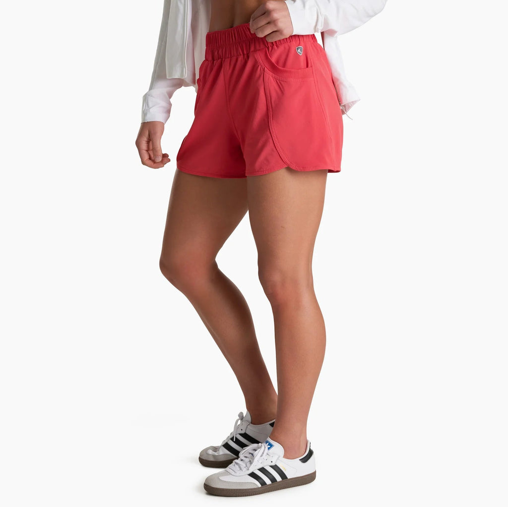 A model wearing bright coral-red athletic shorts with a white shirt and white sneakers, shown from the waist down against a plain white background.