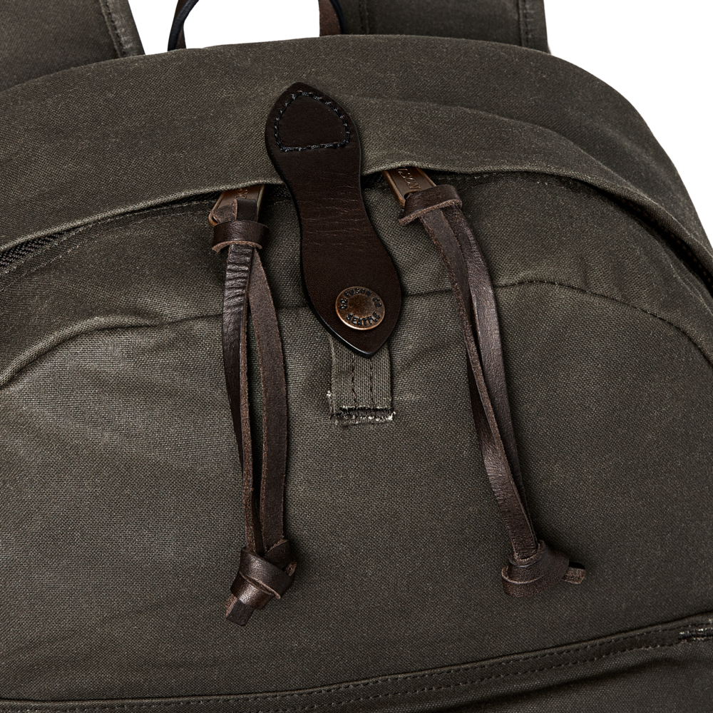 Close-up of an olive green canvas backpack with dark brown leather straps and a metal snap closure, shown against a white background.