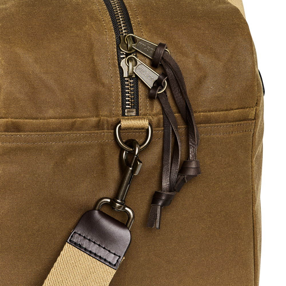Close-up of a brown canvas bag with a black zipper, dark brown leather pulls, metal hardware, and part of a beige-and-brown shoulder strap on a white background.