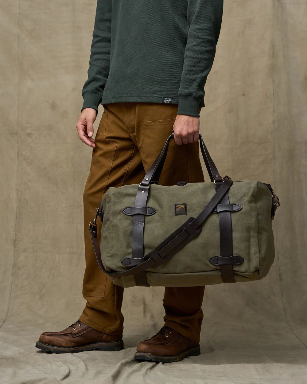 A person wearing a dark green sweater, brown pants, and brown boots carries an olive green duffel bag with dark brown leather straps against a neutral beige backdrop.