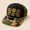 A black and camouflage trucker cap with yellow embroidered text and a sun logo on the front, set against a solid beige background.