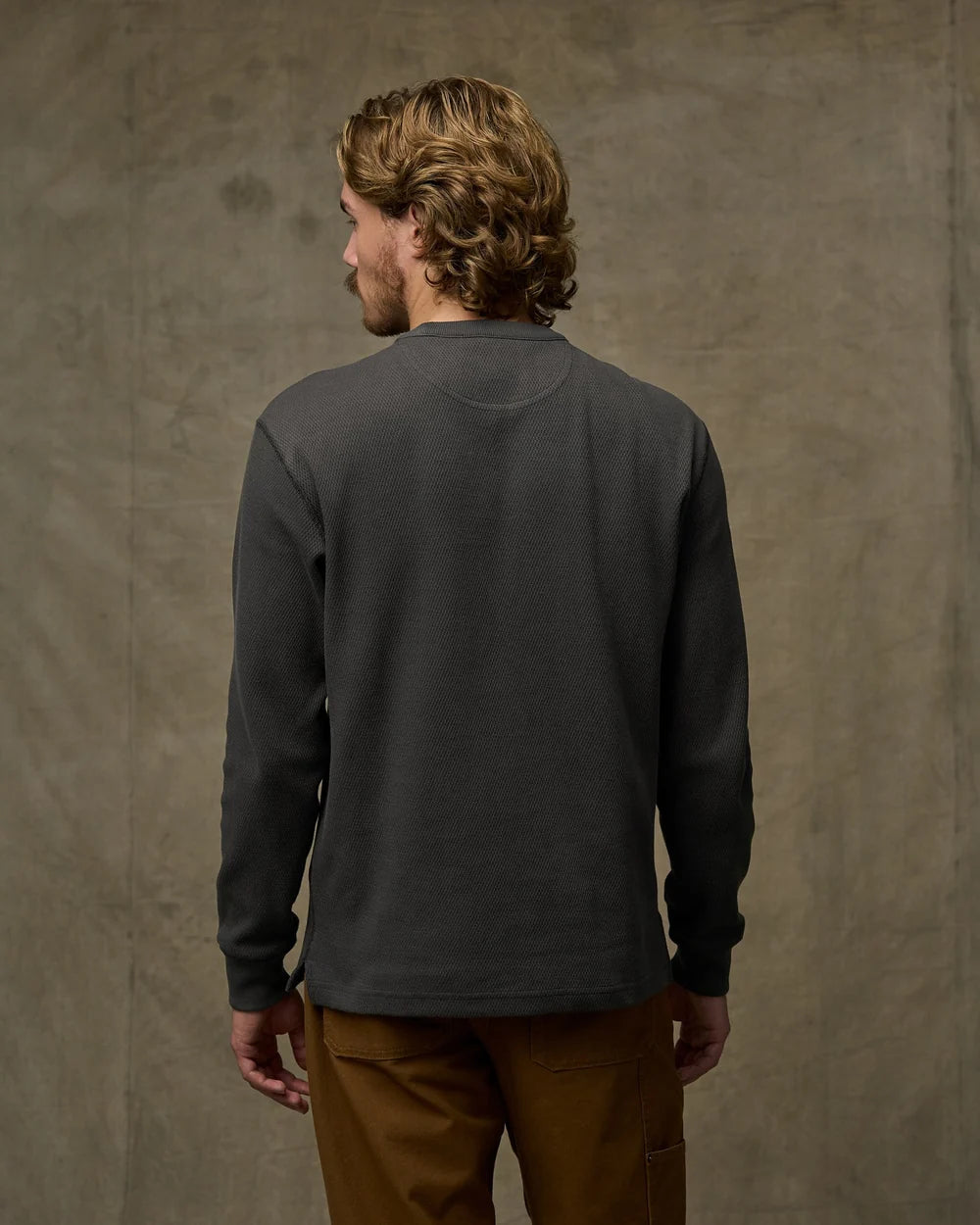 A man with wavy light brown hair is shown from behind wearing a dark charcoal gray long-sleeve crewneck shirt and brown pants, standing against a textured gray concrete wall.