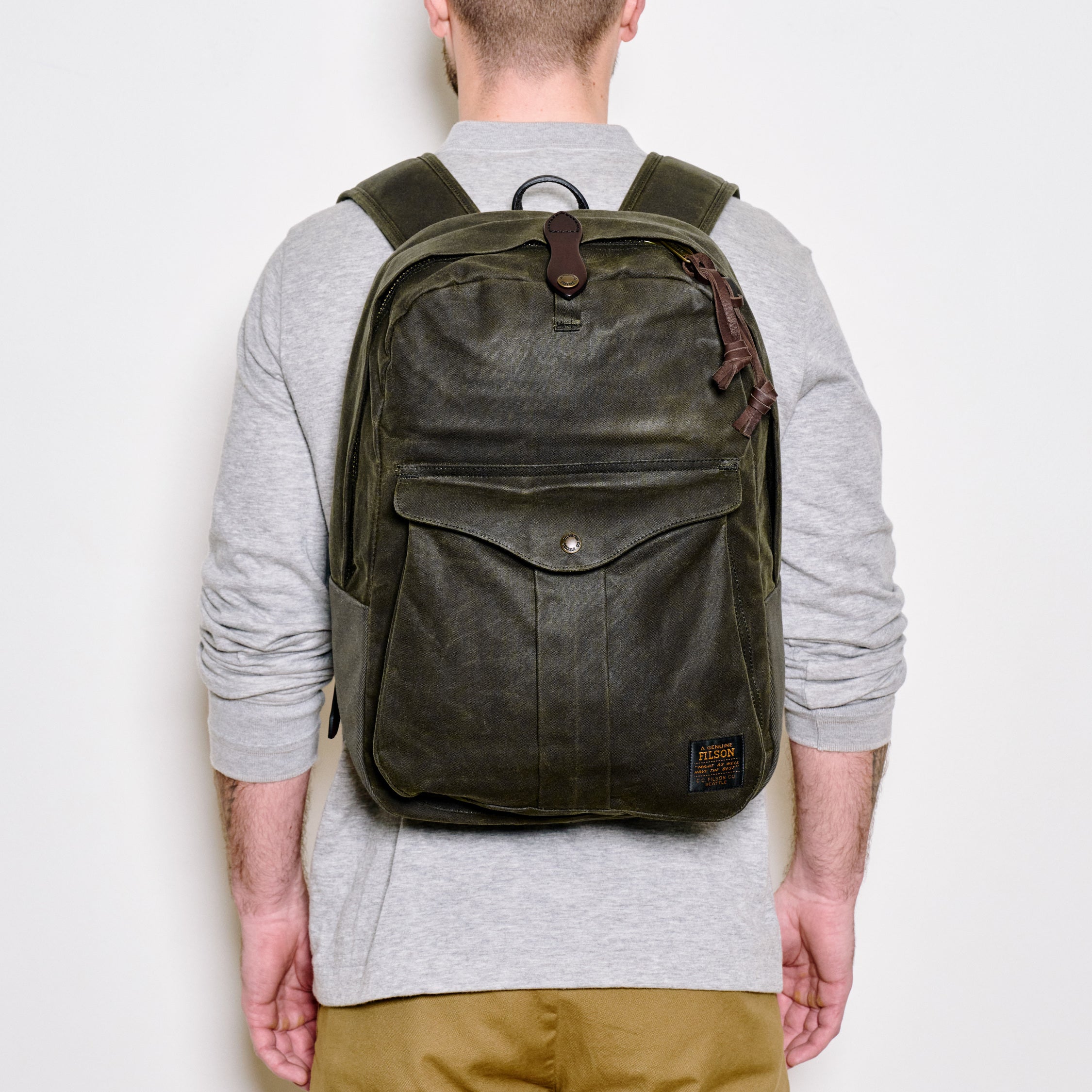 A man viewed from behind wearing an olive green waxed canvas backpack with brown leather accents, over a light gray long-sleeve shirt and khaki pants, against a plain white background.