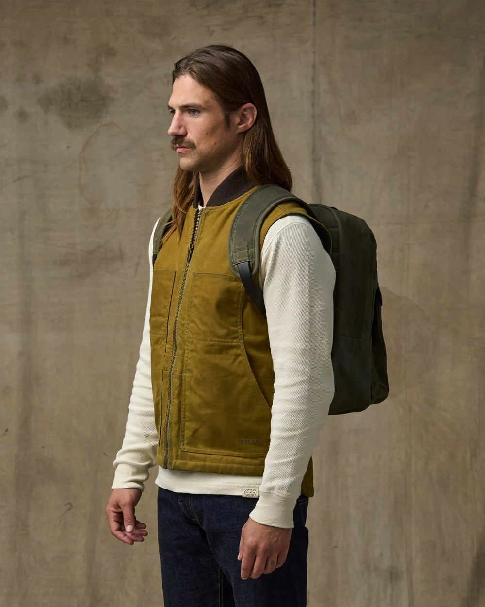 A man with long brown hair and a mustache stands in profile wearing a mustard yellow zip-front vest over a cream long-sleeve shirt, with a dark green backpack, against a textured concrete wall.