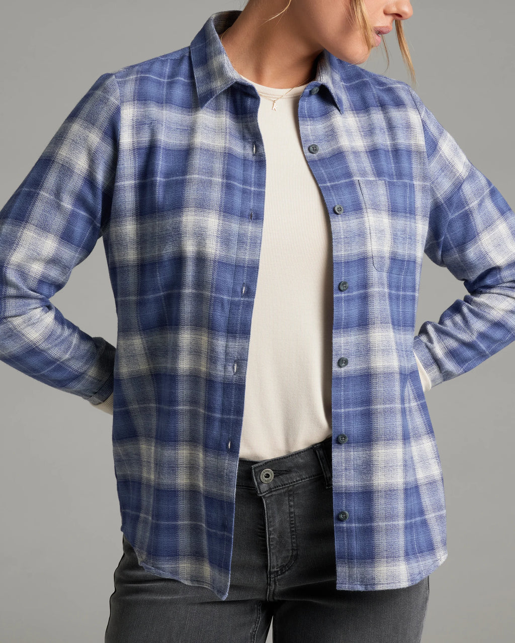 A woman wearing a blue and white plaid button-up shirt over a cream ribbed top and dark gray jeans, shown from the neck down against a solid gray studio background.