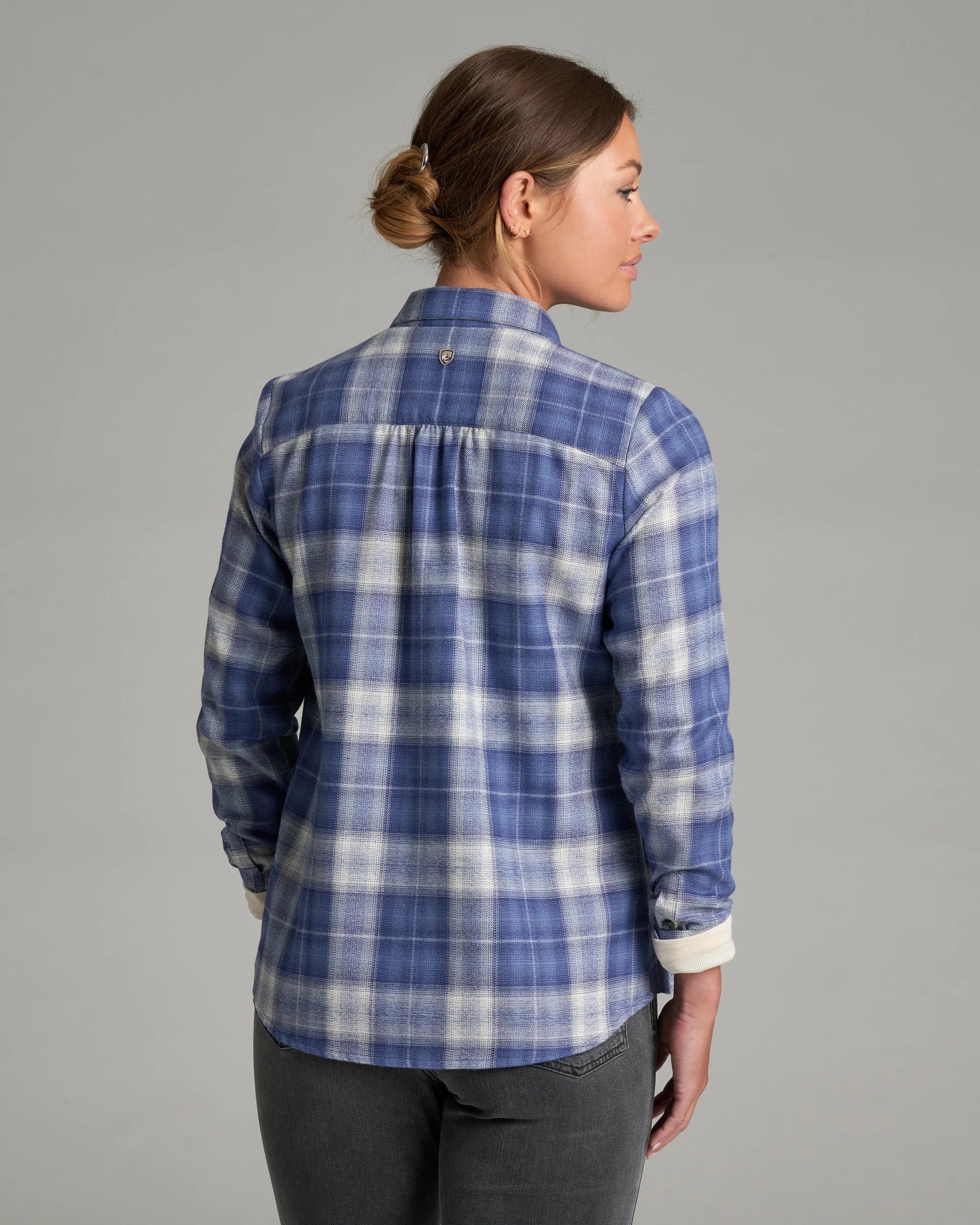A woman shown in side profile wearing a blue and white plaid button-up shirt with dark gray jeans, photographed against a plain light gray studio background.