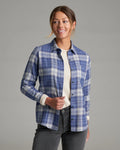 A woman modeling a blue and white plaid button-up shirt over a cream top and dark gray jeans, photographed against a solid gray studio background.