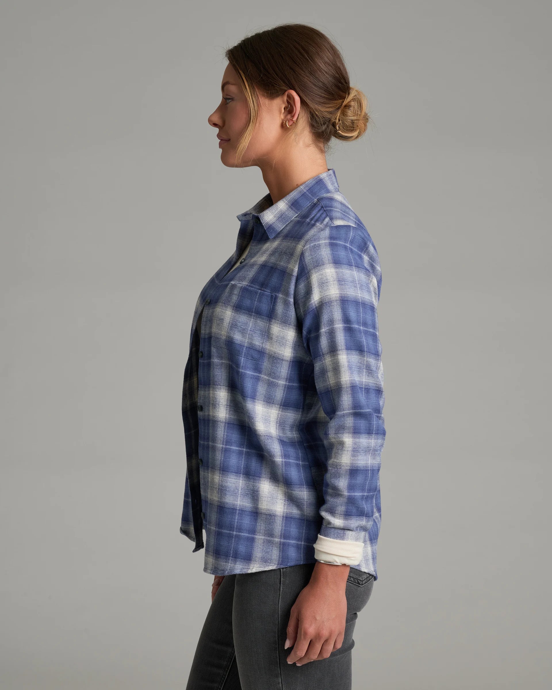 A woman shown in side profile wearing a blue and white plaid button-up shirt with dark gray jeans, photographed against a plain light gray studio background.