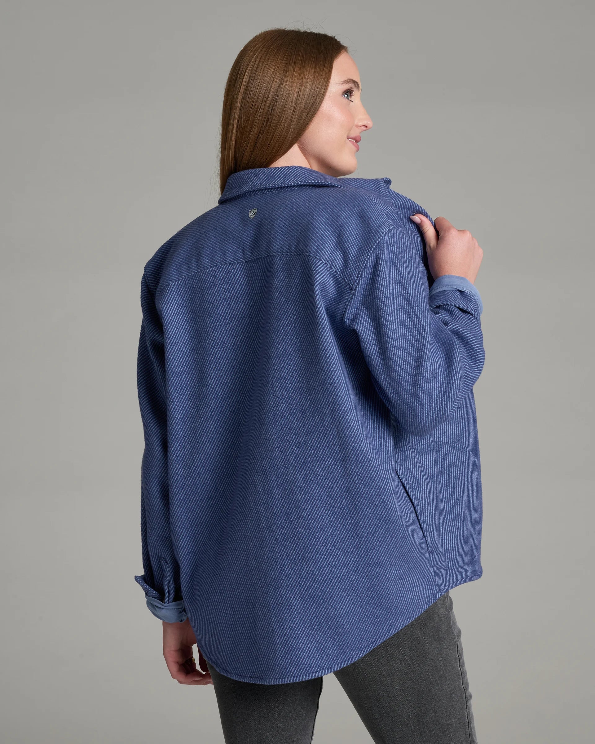 A woman wearing a textured blue button-up shirt jacket, shown from a three-quarters back view against a solid gray studio background.