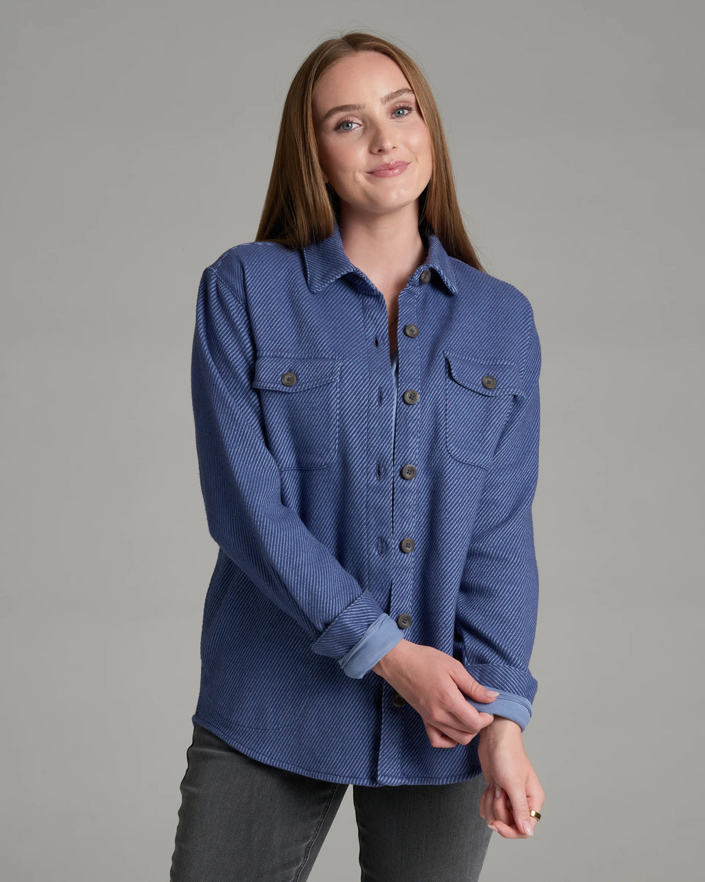 A woman modeling a textured blue button-up shirt with two chest pockets and dark buttons, paired with gray pants, against a solid gray studio background.