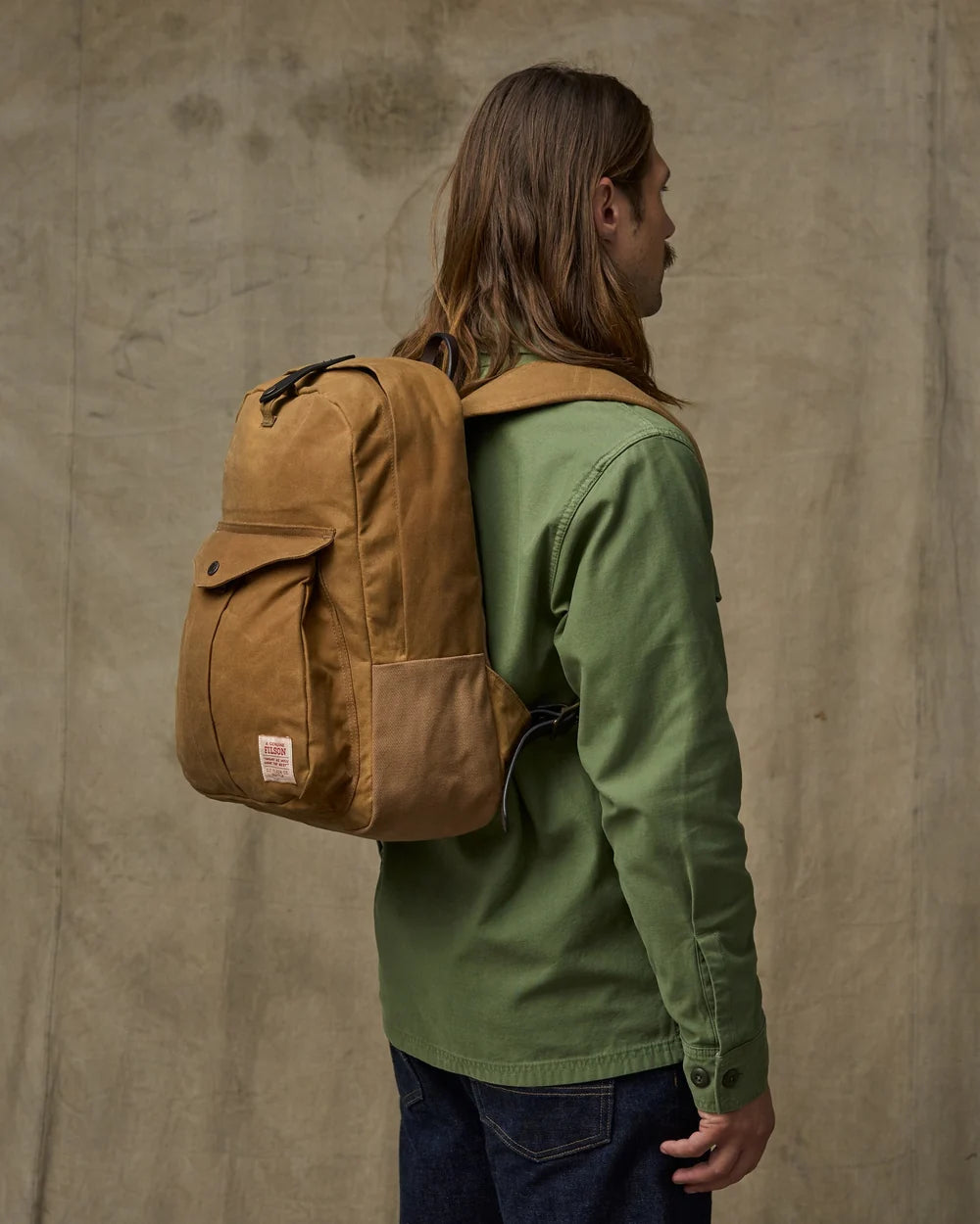 Person wearing a brown backpack and green jacket against a beige wall