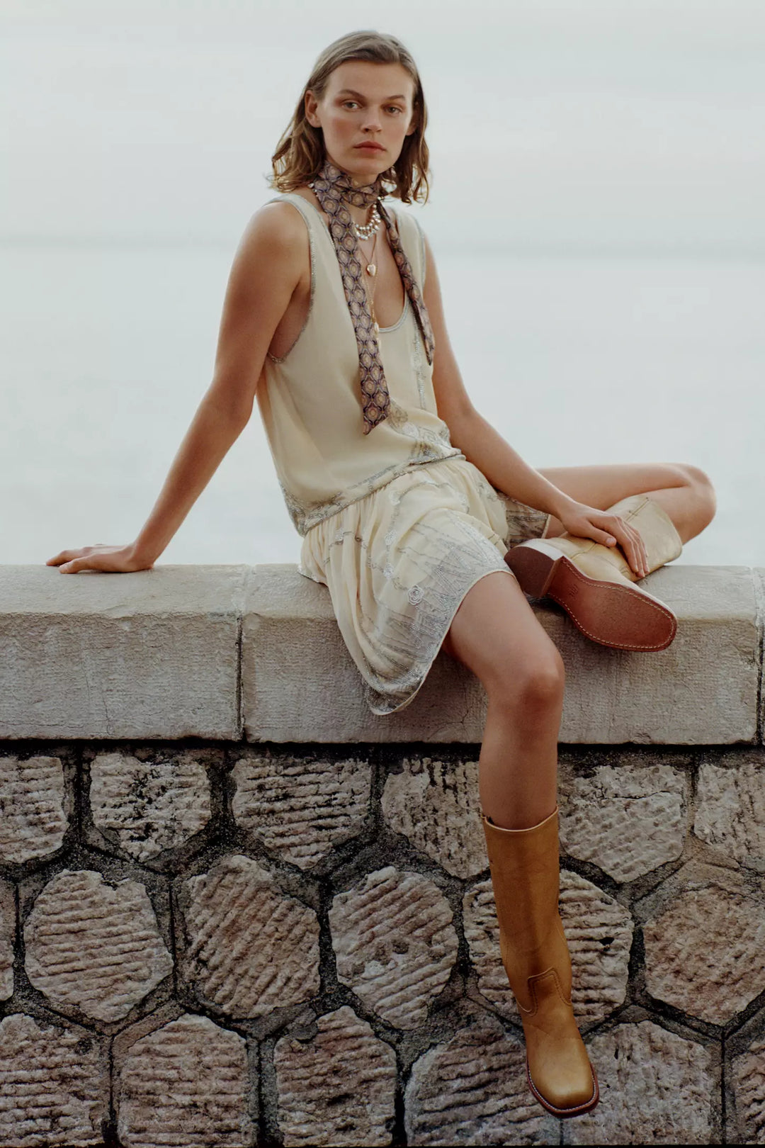 Woman in a light dress and brown boots sitting on a stone wall with a neutral background