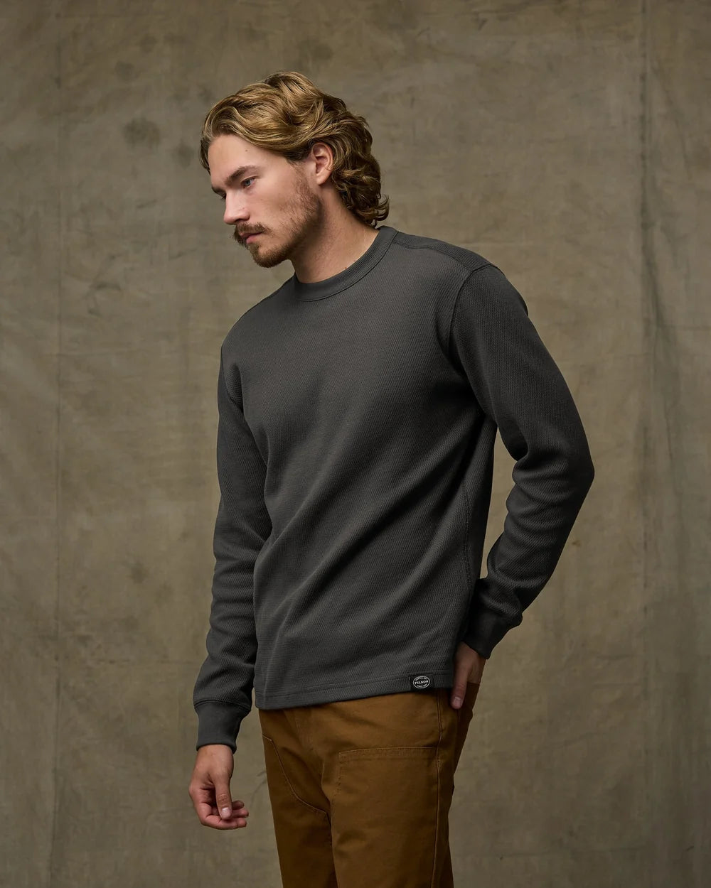 Man wearing a dark gray sweatshirt and brown pants against a textured beige background