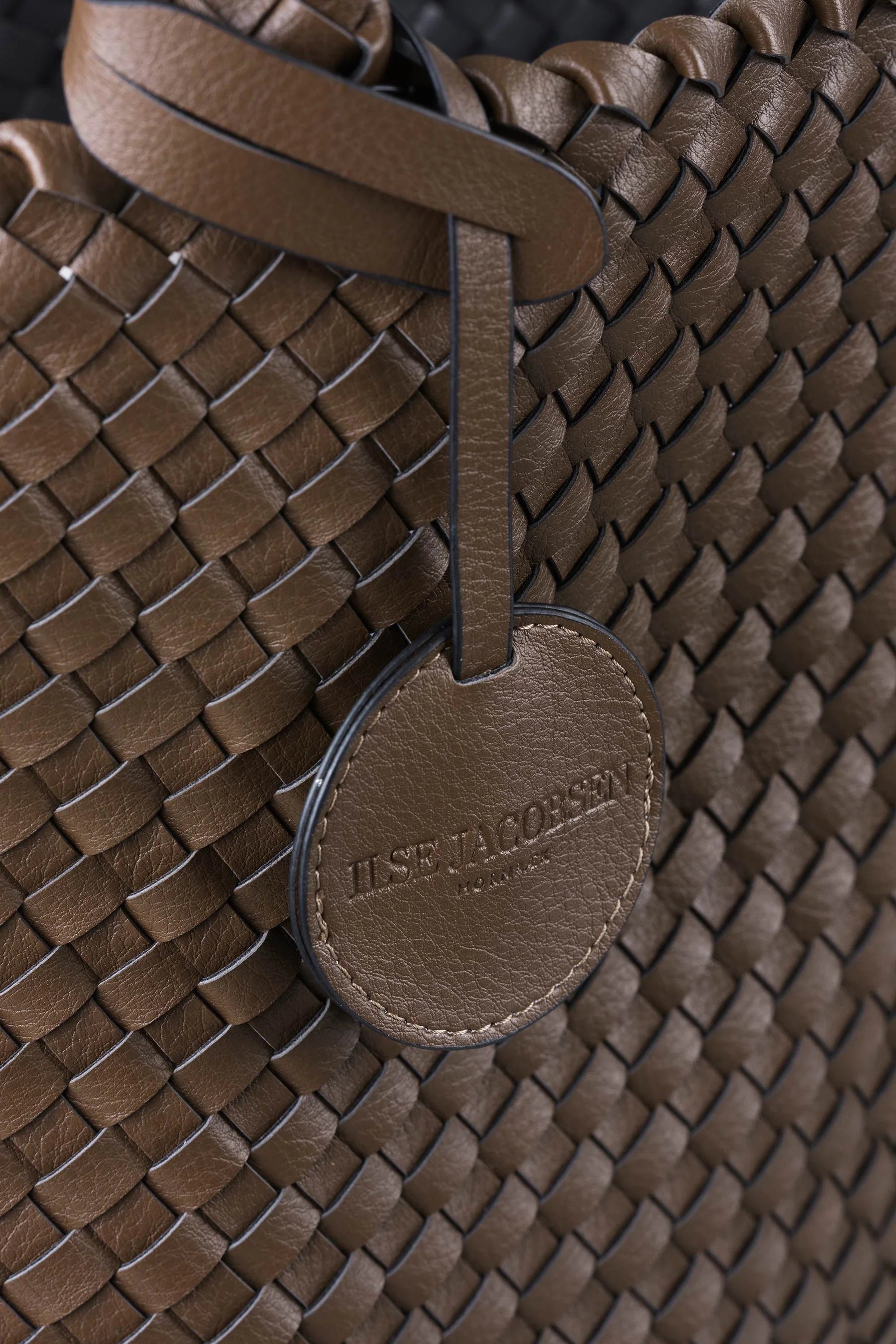 Close-up of a brown woven leather handbag with a round leather brand tag reading 'ILSE JACOPSSEN PARIS' hanging from a strap.