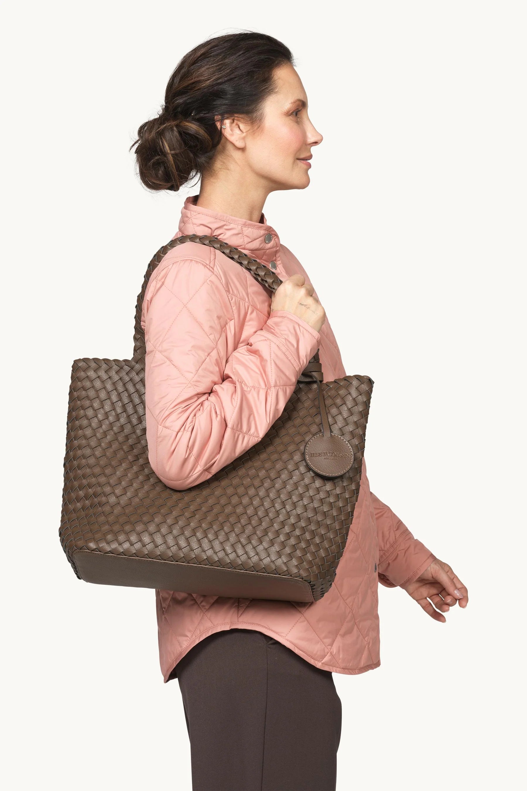 A woman in a pink quilted jacket and dark pants carries a large brown woven leather tote bag over her shoulder, shown in profile against a plain light background.