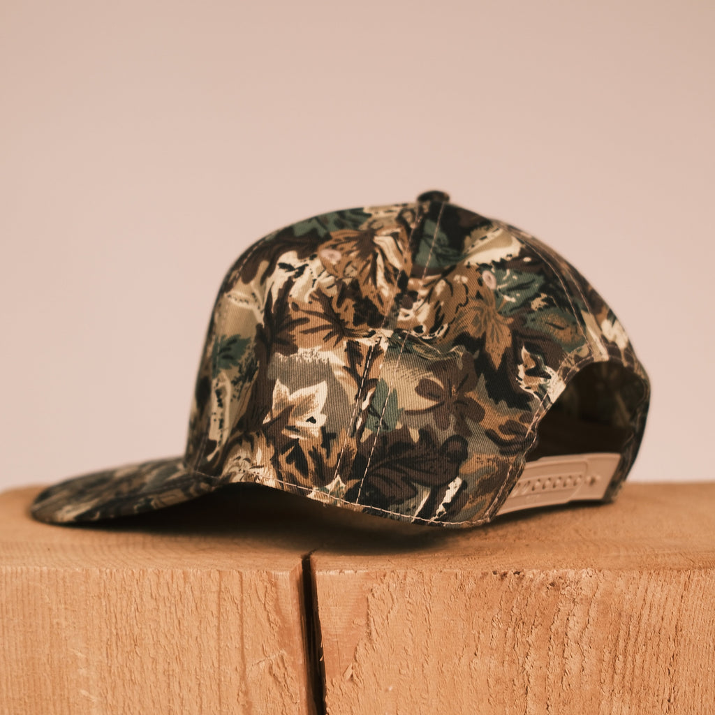A camouflage baseball cap with a floral pattern, hanging from a wooden stand against a solid beige background.