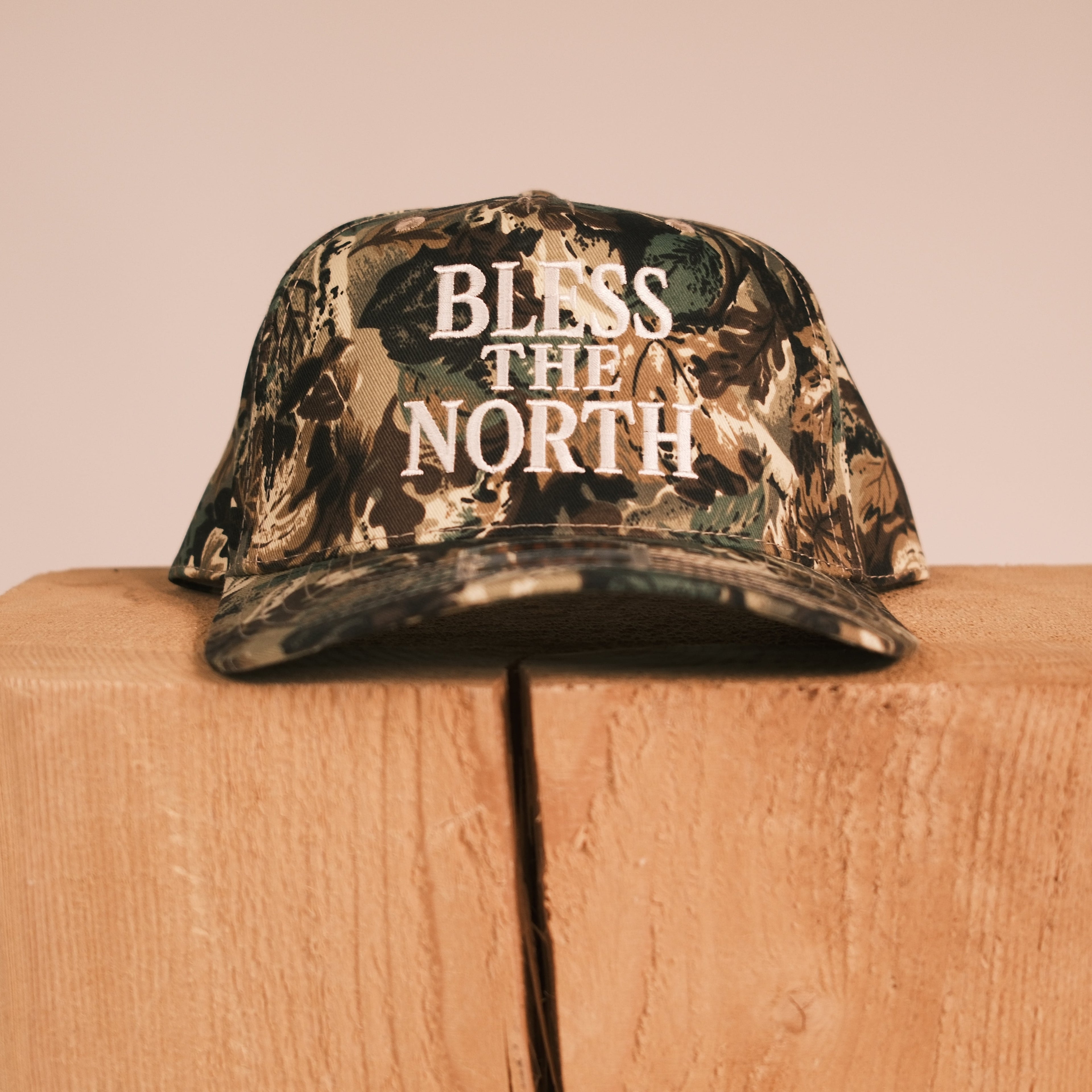 A camouflage baseball cap with 'BLESS THE NORTH' printed in white text on the front, displayed on top of a wooden block against a plain beige background.