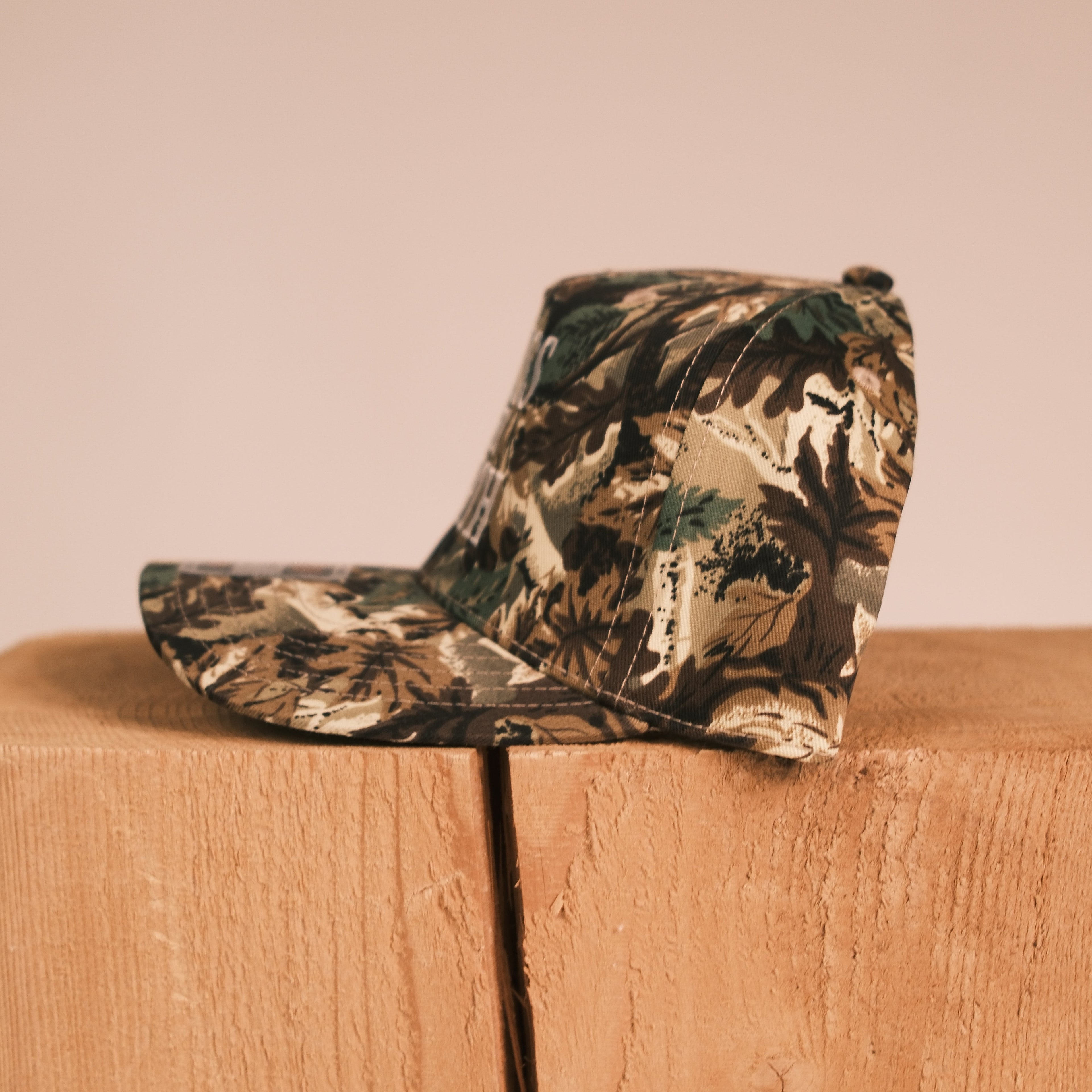 A camouflage baseball cap resting on a wooden block against a solid beige background.